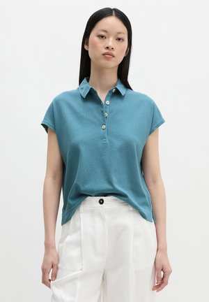 Vrouw met lang zwart haar die een turquoise korte mouwen overhemd met kraag en witte hoge taille broek draagt, staande tegen een effen achtergrond.