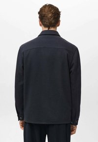 Camicetta giacca in lana blu navy con una vestibilità rilassata, polsini a bottoni e colletto piatto, caratterizzata da una superficie liscia e un design minimalista.