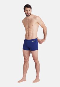 Marineblaue Badshorts aus dehnbarem Material, mit einem Logo am Bund. Das Design ist eng geschnitten und reicht bis zur Mitte des Oberschenkels.