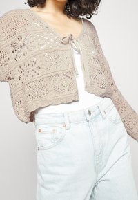 Cardigan corto de punto en color beige claro con un patrón calado, que presenta un cierre con lazo en el frente. Combinado con jeans de tiro alto en color azul claro.