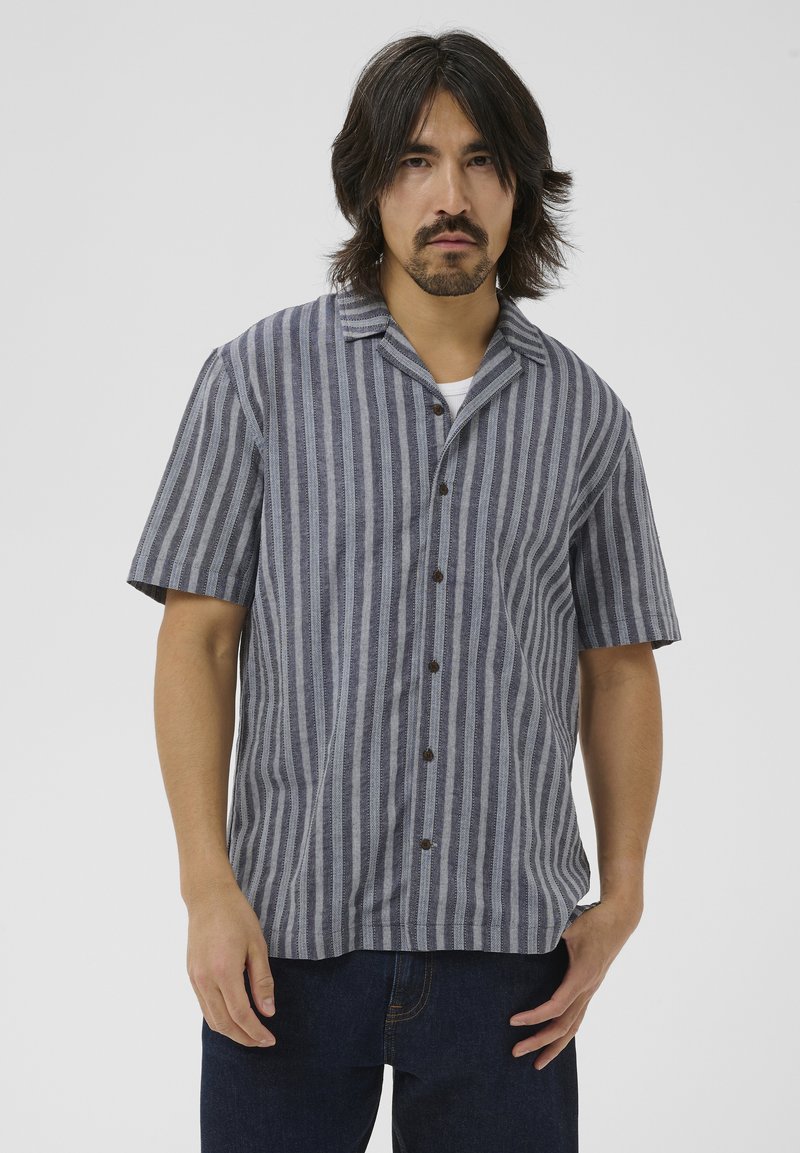 Homme aux cheveux mi-longs portant une chemise à manches courtes à rayures verticales grises et bleu marine et un jean foncé, debout devant un fond uni.
