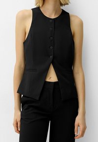 Kvinna klädd i en ärmlös svart väst med knappar och framficka, ihop med svarta skräddarsydda byxor mot en enkel ljus bakgrund.