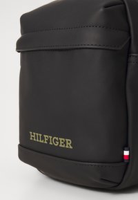 Borsa in tessuto sintetico nero e liscio con una patta frontale, caratterizzata da un logo "HILFIGER" in verde oliva e un piccolo accento tricolore sul lato.