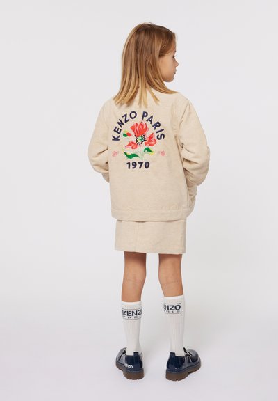 Veste en velours côtelé beige avec un design floral brodé et le texte "KENZO PARIS 1970". Portée avec une jupe assortie, des chaussettes blanches et des chaussures de couleur marine.