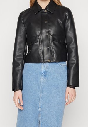 Femme portant une veste courte en cuir noir avec boutons-pression et poches à rabat, associée à une jupe en jean bleu clair.