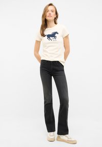 Camiseta de algodón color crema con un gráfico de un caballo en navy, combinada con jeans acampanados negros y zapatillas beige con toques en crema.