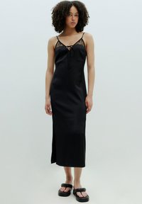 Zwarte satijnen slipdress met kantdetails, een strik aan de voorkant, verstelbare bandjes en een zijsplit, gecombineerd met chunky zwarte sandalen.