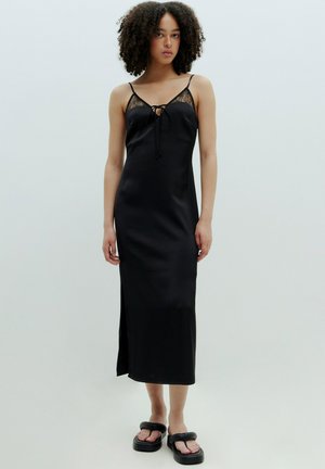 Schwarzes Satin-Nachtkleid mit Spitzenrandeingenschaften, einer vorderen Bindung, verstellbaren Trägern und einem Seitenschlitz, kombiniert mit klobigen schwarzen Sandalen.