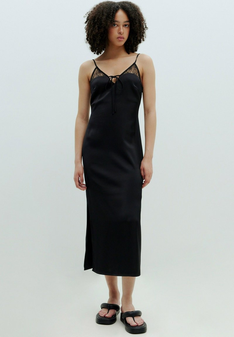 Zwarte satijnen slipdress met kantdetails, een strik aan de voorkant, verstelbare bandjes en een zijsplit, gecombineerd met chunky zwarte sandalen.