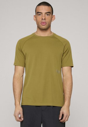 Mann trägt ein olivgrünes Sport-T-Shirt mit kurzen Ärmeln und schwarze Shorts, steht vor einem schlichten hellgrauen Hintergrund.