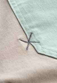 Textielpatch in mintgroene en beige stoffen, met een grijze geborduurde X-detail en zichtbare stiksels langs de randen.