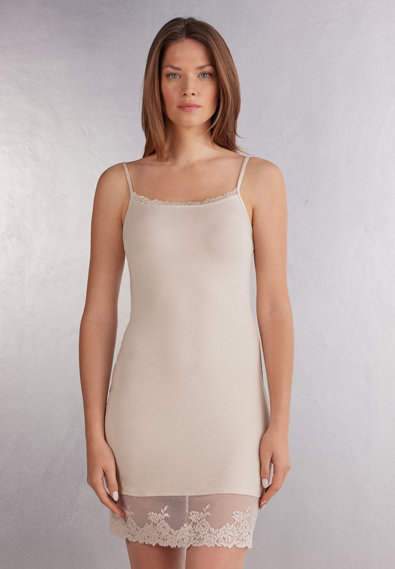 Beige glidklänning i slät tyg med spetsdetaljer vid halsringningen och fållen. Har tunna justerbara axelband och en figursydd silhuett.