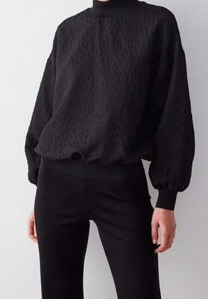 Nainen pukeutunut mustaan teksturoituun pitkähihainen puseroon, jossa on pullistetut hihat ja korkea kaulus, yhdistettynä mustiin istuviin housuihin.