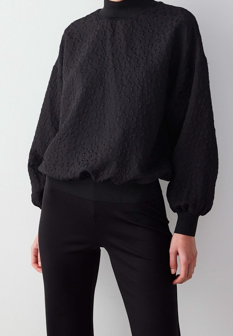 Femme portant un haut noir texturé à manches longues bouffantes et col montant, associé à un pantalon noir ajusté.
