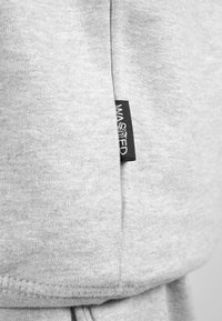 Harmaa kangas, jossa on pehmeä tekstuuri, ja sivussa etiketti, jossa lukee "WASTED" valkoisella mustalla taustalla. Reunassa näkyy ompelusauma.