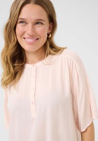 Cămașă roz deschis, cu mâneci scurte, textură moale și plisuri subtile la umeri. Prezintă un guler rotund și cinci butoane asortate.