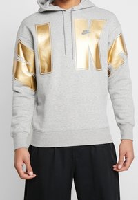 Grå huvtröja med stort text i guldfolie "NIKE" och detaljer på ärmarna. Tillverkad av ett mjukt material med ribbade ärmslut.