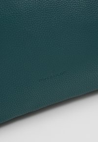 Surface en cuir texturée bleu sarcelle foncé avec un marquage en relief discret « Tom Tailor » près du bord inférieur.
