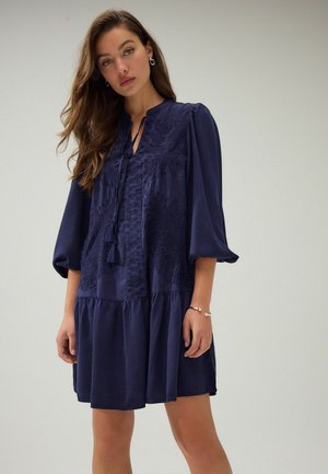 Navy blue dress with a tiered skirt, featuring a textured lace panel and puff sleeves. Ties at the neckline, crafted from a smooth fabric.