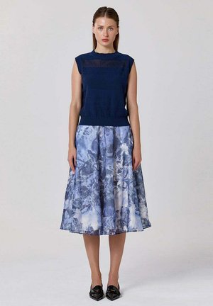 Femme portant un haut sans manches bleu marine et une jupe midi fleurie bleue, debout les bras détendus le long du corps devant un fond uni.