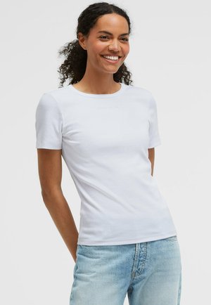 Lächelnde Frau, die ein schlichtes weißes kurzärmliges Shirt und hellblaue Jeans trägt und mit den Händen hinter dem Rücken vor einem weißen Hintergrund steht.