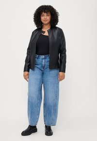 Zwarte neplederen jas met een ronde kraag, gecombineerd met lichtblauwe high-waisted jeans en zwarte enkellaarsjes. Krullend haar zichtbaar.
