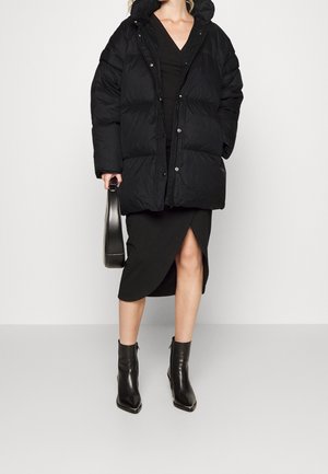 Mujer con un abrigo acolchado negro sobre un vestido negro de cuello en V con una abertura al frente, llevando un bolso negro y botas negras hasta el tobillo.