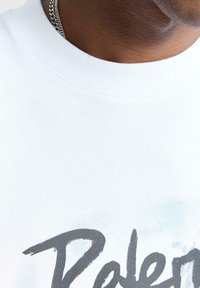 Valkoinen crewneck-huppari, jossa on teksturoitu pinta ja suuri harmaa graafinen painatus. Näkyvät saumat ja kauluksen yksityiskohta.