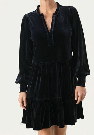 Femme portant une robe noire en velours à manches longues avec un col à volants et une jupe à volants échelonnés, debout devant un fond uni.