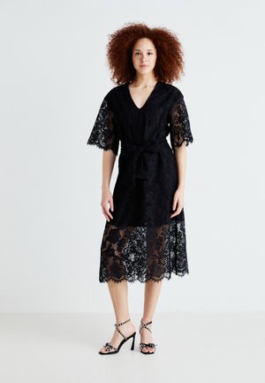 Black lace dress with a v-neck, short sleeves, and a tie belt. Features a sheer lace hem and intricate floral patterns. Paired with black heeled sandals.