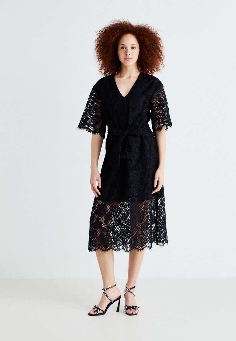Robe en dentelle noire avec un décolleté en V, des manches courtes et une ceinture à nouer. Présente un ourlet en dentelle transparente et des motifs floraux complexes. Associée à des sandales à talons noires.
