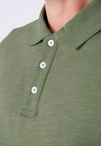 Maslinasto zelena polo majica od mekanog tkanja; ima ovratnik, tri bijela gumba i suptilno teksturiranu površinu. Jednostavan dizajn bez uzoraka.