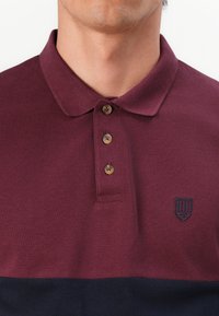Burgundy-polkka paita, jossa on teksturoitu pinta, klassinen kaulus, kolmonen napitus ja tummansininen alaosa, jossa on kirjailtu emblem.