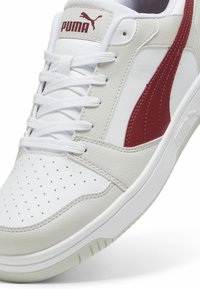Baskets blanches Puma avec bout perforé, lacets blancs, logo Puma rouge sur la languette et bande rouge sur le côté.