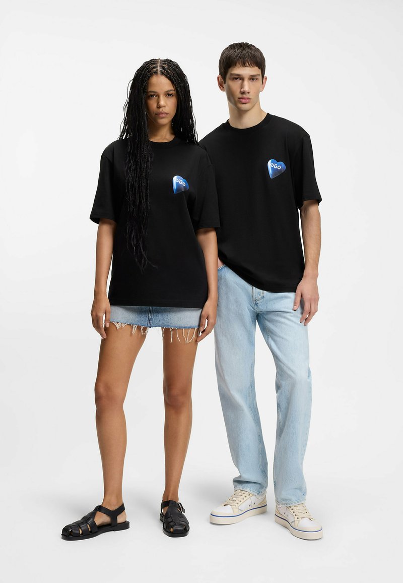 Jeune femme et homme debout côte à côte, portant des t-shirts noirs avec des logos de cœur bleus, jupe en jean, jean clair et chaussures décontractées sur fond blanc.