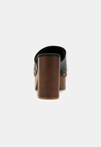 Zoccolo nero con tacco alto e design arrotondato, caratterizzato da un spesso tacco in legno marrone e borchie dorate decorative. Texture liscia in tutto il prodotto.