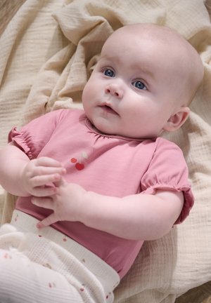 Baby met blauwe ogen in een roze shirt met kersenprint, liggend op een beige deken, handen zachtjes samengevouwen.