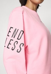 Sweatshirt rose à manches longues présentant un texte noir portant l'inscription « ENDLESS » sur le bras gauche. Le matériau semble doux et texturé.