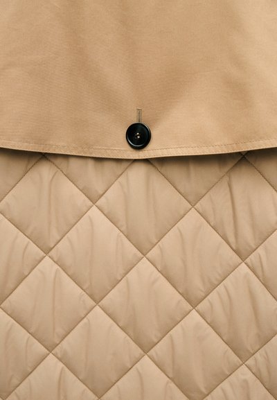 Μπεζ υφασμάτινο πάπλωμα κάτω από λείο μπεζ ύφασμα με καπάκι που στερεώνεται με ένα μαύρο κουμπί.