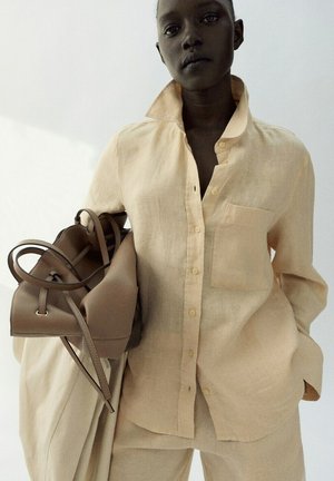 Persona de piel oscura que lleva una camisa y pantalones de lino beige, sosteniendo un bolso de cuero topo y un abrigo a juego contra un fondo claro y liso.