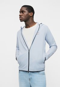 Άνδρας που φοράει ανοιχτό μπλε μπουφάν με φερμουάρ πάνω από άσπρο πουκάμισο και ανοιχτό μπλε τζιν, στέκεται με τα χέρια στις τσέπες, κοιτώντας προς τα αριστερά.