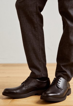 Schwarze Lederhalbschuhe mit polierter Oberfläche, runden Zehen und farblich abgestimmten Schuhbändern, kombiniert mit grauen Nadelstreifen-Hosen, auf einem Holzboden.