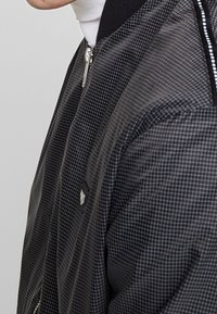 Svart bomberjacka med rutmönster, som har dragkedjestängning och ribbad krage. Tyget har en slät textur och en minimalistisk design.