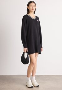 Μαύρο oversized φόρεμα με V-λαιμόκοψη και λευκές ρίγες, διαθέτει μικρό λογότυπο. Φορεμένο με λευκές κάλτσες και χοντροπάτωνα αθλητικά παπούτσια.