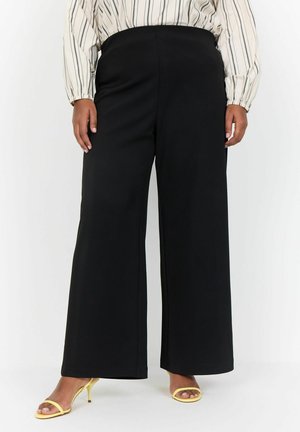 Schwarze, weite Hosen mit elastischem Bund, aus einem glatten, weichen Stoff. Kombiniert mit gelben, hochhackigen Sandalen. Vertikale Streifen auf der Bluse.