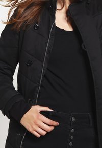 Svart vadderad jacka med tryckknappar, frontdragkedja och elastiska ärmar, worn över en svart topp och svart kjol med knapppar.