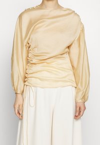 Pitkähihaista paita vaaleankeltaisesta satiinista, jossa on kootut sivut ja pyöreä pääntie, jossa on napituskoristeet olkapäällä. Sujuva pinta.
