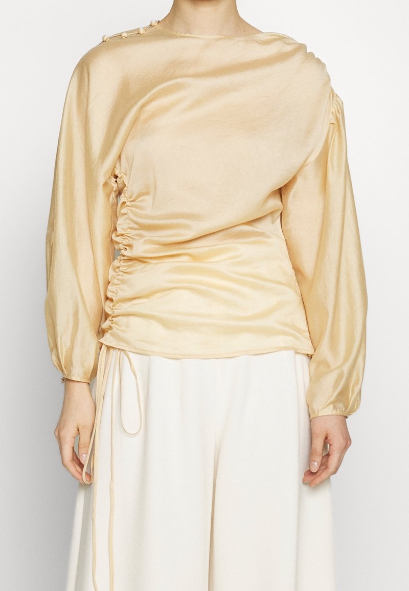 Pitkähihaista paita vaaleankeltaisesta satiinista, jossa on kootut sivut ja pyöreä pääntie, jossa on napituskoristeet olkapäällä. Sujuva pinta.