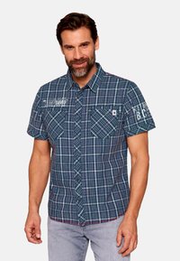 Camicia a manica corta a quadri blu navy e verde, con due tasche sul petto, chiusura con bottoni e testo ricamato sulla spalla. Materiale in cotone.