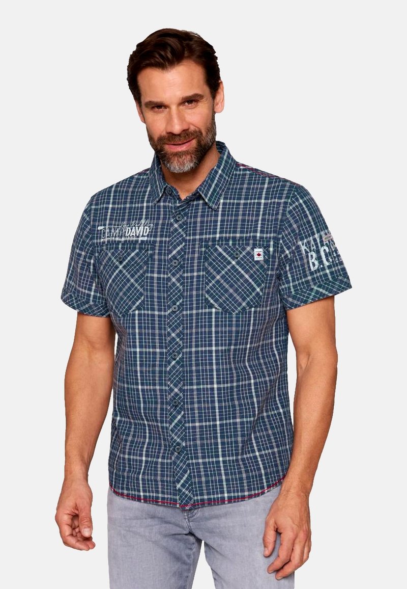 Camicia a manica corta a quadri blu navy e verde, con due tasche sul petto, chiusura con bottoni e testo ricamato sulla spalla. Materiale in cotone.